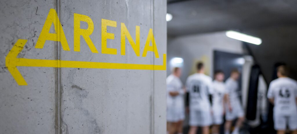 HANDBALL 2024/25: THW Kiel im Trainingslager in Graz Der gelbe Schriftzug ARENA mit einem Pfeil ist auf eine Betonwand gemalt; im verschwommenen Hintergrund steht eine Gruppe von Menschen in weißen Sportuniformen unter Neonlicht zusammen.