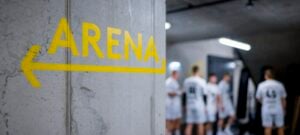 Ein gelber Pfeil mit der Aufschrift ARENA ist auf eine Betonwand gemalt und zeigt nach links. Im Hintergrund ist eine Gruppe von Sportlern in weißen Uniformen zu erkennen.