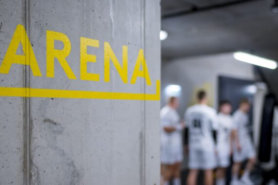 Ein gelber Pfeil mit der Aufschrift ARENA ist auf eine Betonwand gemalt und zeigt nach links. Im Hintergrund ist eine Gruppe von Sportlern in weißen Uniformen zu erkennen.