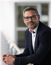 Ein Mann mit kurzen Haaren und Brille, der einen dunklen Anzug und ein weißes Hemd trägt, lehnt in einem Haus an einem Geländer und schaut mit einem leichten Lächeln in die Kamera. Der unscharfe Hintergrund deutet auf eine Umgebung hin, die mit der THW Kiel Geschäftsführung in Verbindung steht.