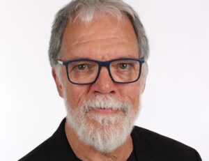 Ein älterer Mann mit grauen Haaren, weißem Vollbart und blau gerahmter Brille, der eine schwarze Jacke trägt, posiert vor einem schlichten weißen Hintergrund - und fängt damit die nachdenkliche Stimmung von Patrick Wienceks Abschied vom THW Kiel nach der Handball-Saison 2025/2026 ein.