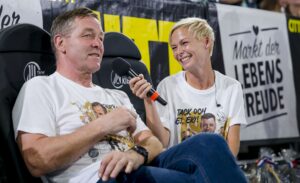 Eine Frau mit einem Mikrofon lächelt, während sie einen sitzenden Mann interviewt, beide in passenden weißen Grafik-T-Shirts. Hinter ihnen prangt ein Banner mit der Aufschrift "Markt der Lebensfreude", das auf die THW Kiel Saison 2025/2026 und Patrick Wienceks Handball Abschied hinweist.