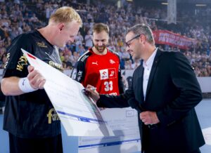 Ein lächelnder Mann in einem Anzug überreicht einem Sportler in einem schwarzen Trikot einen großen Scheck, während ein anderer Sportler in einem roten Trikot in der Nähe steht. Alle befinden sich auf einem Sportplatz, umgeben von einer jubelnden Menge.