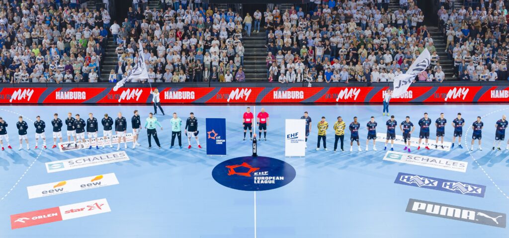HANDBALL 2024/25: THW Kiel - Limoges Handball Professionelle Handballmannschaften stellen sich vor einem Spiel in einer vollbesetzten Arena auf einem blauen Spielfeld auf, mit Schiedsrichtern, Maskottchen und Fahnen, während das Publikum von der Tribüne aus zusieht.