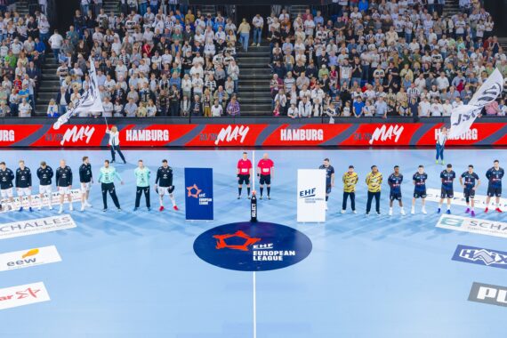 Professionelle Handballmannschaften stellen sich vor einem Spiel in einer vollbesetzten Arena auf einem blauen Spielfeld auf, mit Schiedsrichtern, Maskottchen und Fahnen, während das Publikum von der Tribüne aus zusieht.