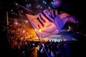 Eine große Menschenmenge verfolgt eine Indoor-Sportveranstaltung mit dramatischer Beleuchtung und einer riesigen wehenden Flagge im Vordergrund. Helle Lichter und Feuereffekte verstärken die energiegeladene Atmosphäre.
