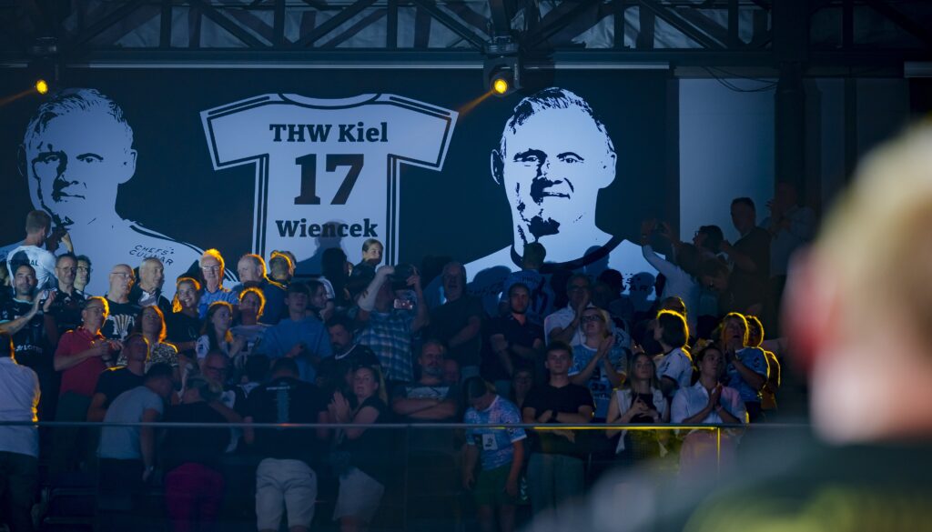 Die Menge steht und applaudiert vor einem großen Banner, auf dem ein Trikot mit dem Namen Wiencek, der Nummer 17 und dem THW Kiel sowie zwei Porträts des Spielers zu sehen sind, in einer schwach beleuchteten Arena.