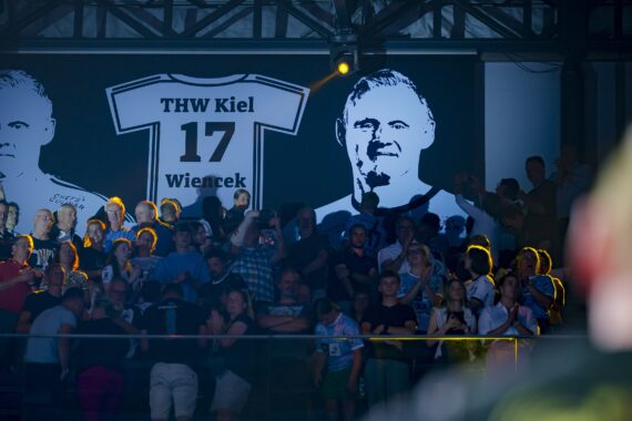 Die Menge steht und applaudiert vor einem großen Banner, auf dem ein Trikot mit dem Namen Wiencek, der Nummer 17 und dem THW Kiel sowie zwei Porträts des Spielers zu sehen sind, in einer schwach beleuchteten Arena.