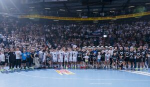 Eine große Gruppe von Handballspielern, Trainern und Betreuern posiert gemeinsam auf einem Hallenplatz mit Blick in die Kamera, während im Hintergrund ein volles Stadion mit jubelnden Fans zu sehen ist.