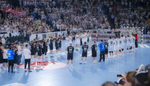 Zwei Handballmannschaften, eine in Schwarz und eine in Weiß, stehen sich in Reihen auf einem blauen Hallenplatz gegenüber, umgeben von einer großen, jubelnden Menge in einer Arena.