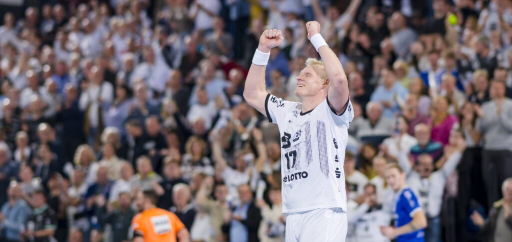 HANDBALL 2024/25: THW Kiel - VfL Gummersbach Ein Handballspieler in einem weißen Trikot jubelt mit erhobenen Armen vor einer jubelnden Menge während eines Hallenspiels. Im Hintergrund sind andere Spieler und Zuschauer zu sehen.