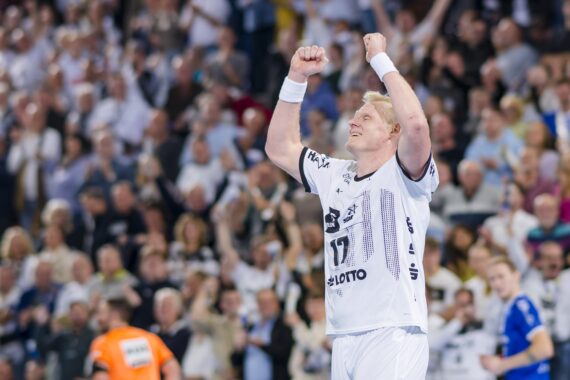 Ein Handballspieler in einem weißen Trikot jubelt mit erhobenen Armen vor einer jubelnden Menge während eines Hallenspiels. Im Hintergrund sind andere Spieler und Zuschauer zu sehen.