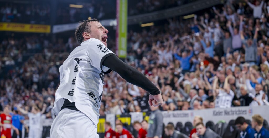 Ein Handballspieler in einem weißen Trikot feiert leidenschaftlich auf dem Spielfeld und schreit mit geballter Faust, während eine große Menschenmenge im Stadion im Hintergrund jubelt.