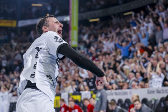 Ein Handballspieler in einem weißen Trikot feiert leidenschaftlich auf dem Spielfeld und schreit mit geballter Faust, während eine große Menschenmenge im Stadion im Hintergrund jubelt.