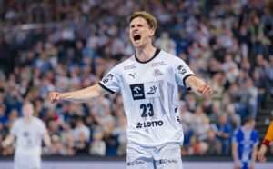 Ein männlicher Handballspieler in einem weißen Trikot mit der Nummer 23 feiert leidenschaftlich mit geballten Fäusten auf einem Spielfeld, umgeben von einer unscharfen Menge und anderen Spielern im Hintergrund.