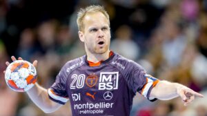 Ein Handballspieler in einem lila Trikot hält während eines Spiels einen Ball in der einen Hand und gestikuliert mit der anderen. Das Trikot trägt die Nummer 20 und verschiedene Sponsorenlogos. Im Hintergrund ist eine unscharfe Menschenmenge zu sehen.