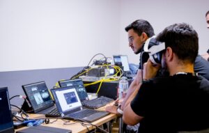 Zwei Männer arbeiten an einem Schreibtisch mit mehreren Laptops und Kabeln; einer schaut auf einen Bildschirm, während der andere ein Virtual-Reality-Headset trägt. Elektronische Geräte und Wasserflaschen stehen auf dem Tisch.
