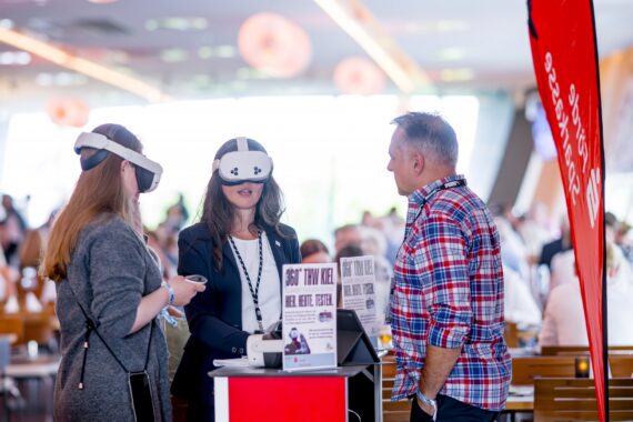 Drei Personen stehen bei einer Veranstaltung um einen kleinen Tisch herum; zwei tragen Virtual-Reality-Headsets, während die dritte Person in einem karierten Hemd zusieht. Der Hintergrund ist hell erleuchtet und mit anderen Teilnehmern gefüllt.