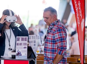 Eine Frau benutzt ein Virtual-Reality-Headset an einem Stand, während ein Mann in einem karierten Hemd in der Nähe steht. Auf dem Tisch liegen Informationsschilder und Broschüren aus. Der Hintergrund ist unscharf mit Menschen und einem roten Banner.