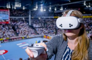 Eine Frau, die ein Virtual-Reality-Headset trägt und einen Controller in der Hand hält, erlebt VR in einem überfüllten Sportstadion, wobei das Publikum und das Spielfeld im Hintergrund verschwimmen.