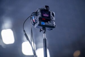 Eine auf einem Stativ montierte Digitalkamera ist nach oben gerichtet und zeigt auf dem Bildschirm eine Bühnen- oder Arenaszene. Die Kamera ist mit Kabeln verbunden, und helle Lichter erzeugen einen unscharfen Hintergrund.