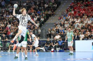 Ein Handballspieler im weiß-schwarz gestreiften Trikot des THW Kiel springt zum Schuss, während ein Abwehrspieler versucht, ihn während des Handball-Bundesliga-Supercups München 2025/2026 zu blocken. Die Füchse Berlin gewinnen vor zahlreichen Zuschauern mit 34:33.