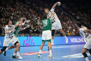 Ein Spieler des THW Kiel in Weiß springt während des Handball-Bundesliga-Supercups 2025/2026 in München zum Schuss, während drei Abwehrspieler der Füchse Berlin ihn abblocken und die zahlreichen Zuschauer ein spannendes 33:34-Spiel verfolgen.