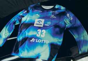 Ein langärmeliges Sporttrikot mit abstraktem Muster in Blau und Petrol, weißem Text und Logos, der Nummer 33 auf der Vorderseite und verschiedenen Sponsorenlogos wie Puma, ORLEN, Star und LOTTO.