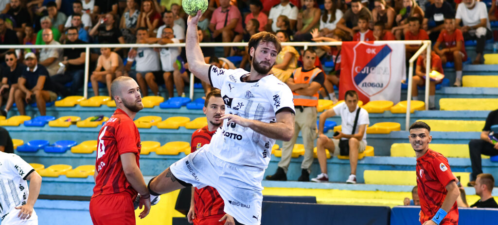 Ein Handballspieler in einem weißen Trikot springt, um den Ball zu werfen, während Spieler in Rot ihn verteidigen. Das Spiel findet in einer Halle statt, während die Zuschauer im Hintergrund von der Tribüne aus zusehen.