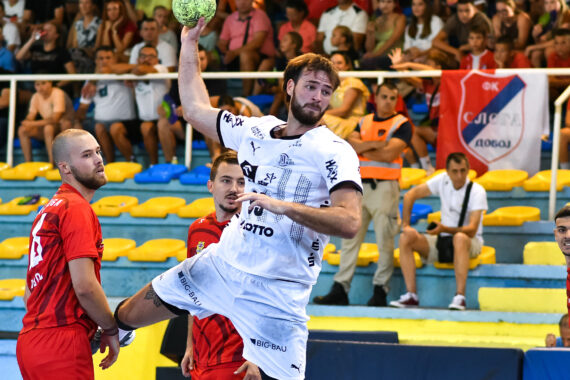 Ein Handballspieler in einem weißen Trikot springt, um den Ball zu werfen, während Spieler in Rot ihn verteidigen. Das Spiel findet in einer Halle statt, während die Zuschauer im Hintergrund von der Tribüne aus zusehen.