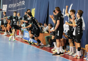 Eine Herren-Handballmannschaft in schwarzen Trikots feiert während eines Spiels am Spielfeldrand, wobei die Spieler klatschen, jubeln und einer auf das Spielfeld läuft.