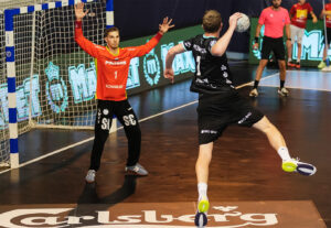 Ein Handballspieler in einem schwarzen Trikot springt zum Schuss, während ein Torwart in einem orangefarbenen Trikot sich darauf vorbereitet, vor dem Tor zu blocken. Andere Spieler und Schiedsrichter sind im Hintergrund zu sehen.