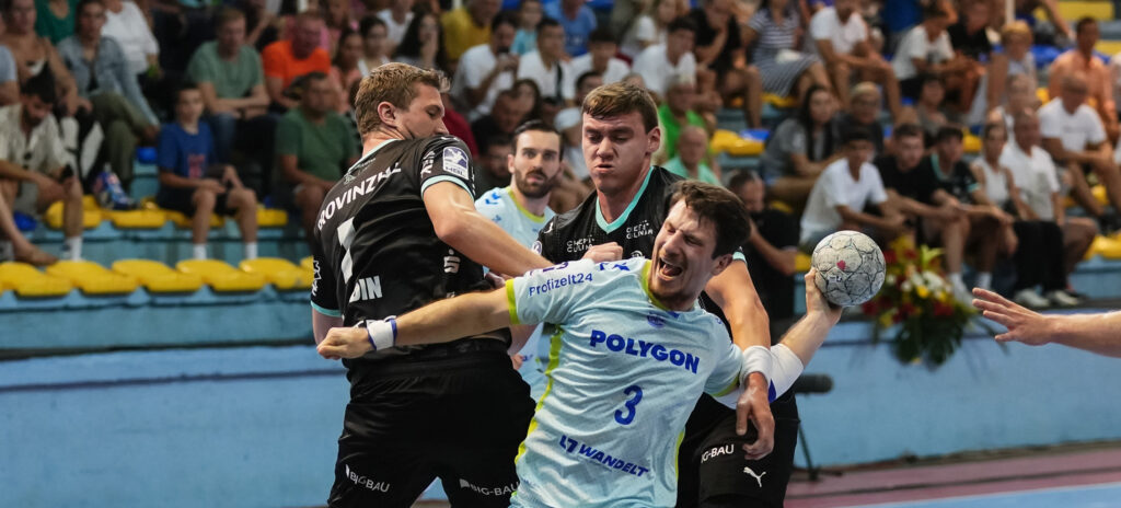Zwei männliche Handballspieler in schwarzen Trikots blockieren einen Spieler in einem hellblauen Trikot, der den Ball hält, während eines intensiven Spiels, bei dem die Zuschauer von gelben und blauen Tribünen im Hintergrund zusehen.