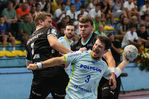 Zwei männliche Handballspieler in schwarzen Trikots blockieren einen Spieler in einem hellblauen Trikot, der den Ball hält, während eines intensiven Spiels, bei dem die Zuschauer von gelben und blauen Tribünen im Hintergrund zusehen.