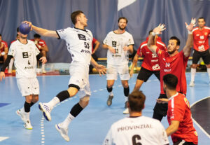 Ein männlicher Handballspieler in einer weißen Uniform springt, um den Ball zu werfen, während Spieler in roten Uniformen den Ball verteidigen. Im Hintergrund sind andere Spieler und ein blaues Spielfeld zu sehen.