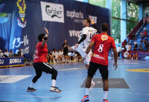 Ein Handballspieler in einem weißen Trikot springt zum Wurf, während zwei Verteidiger in roten Trikots versuchen, ihn während eines Spiels in einer Halle zu blockieren. Die Zuschauer sitzen im Hintergrund.