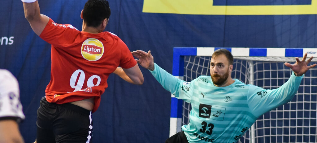 Ein männlicher Handballspieler in einem roten Trikot mit der Nummer 98 springt und schießt auf das Tor, während ein männlicher Torwart in einem hellblauen Trikot mit der Nummer 33 die Arme hochreißt, um den Schuss abzuwehren.