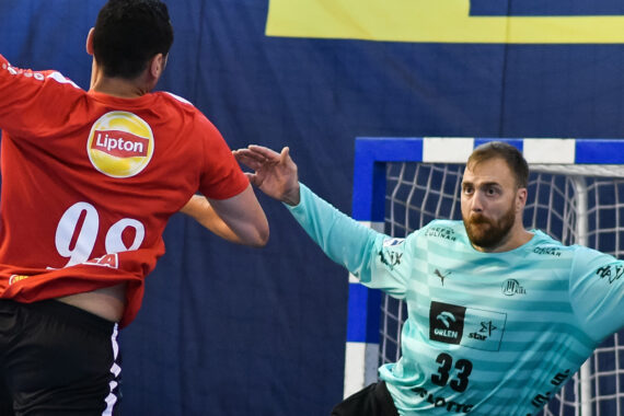 Ein männlicher Handballspieler in einem roten Trikot mit der Nummer 98 springt und schießt auf das Tor, während ein männlicher Torwart in einem hellblauen Trikot mit der Nummer 33 die Arme hochreißt, um den Schuss abzuwehren.