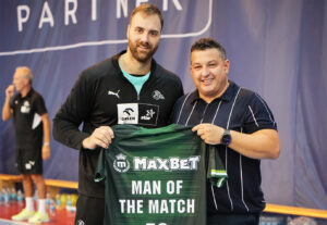 Zwei Männer stehen drinnen und lächeln in die Kamera. Einer hält ein grünes Sporttrikot, auf dem in weißen Buchstaben MAXBET MAN OF THE MATCH steht. Im Hintergrund sind eine verschwommene Sportstätte und Menschen zu sehen.