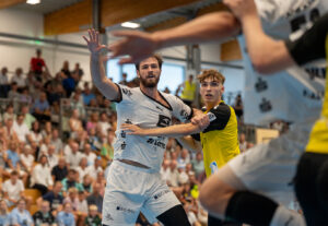 Zwei männliche Handballspieler, einer in einem weißen und einer in einem gelben Trikot, bewachen sich gegenseitig während eines Spiels. Die Zuschauer auf der Tribüne im Hintergrund schauen aufmerksam zu.
