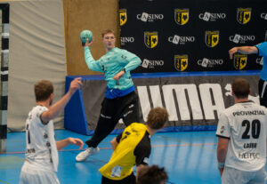 Ein Handballtorwart in einem blauen Trikot bereitet sich auf den Wurf vor, während ein Spieler in einem weißen und ein Spieler in einem gelben Trikot ihm während eines Spiels in einer blauen Halle gegenüberstehen.