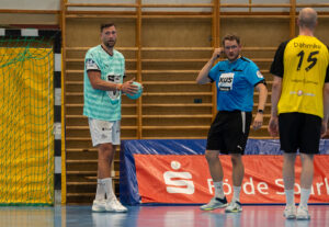 Ein Handballspieler in einem blauen Trikot hält den Ball, während ein Schiedsrichter in einem blauen Hemd ein Zeichen gibt. Ein anderer Spieler in einem gelben Trikot mit der Dahmke 15 auf dem Rücken steht in der Nähe. Im Hintergrund ist eine hölzerne Turnhallenbestuhlung zu sehen.