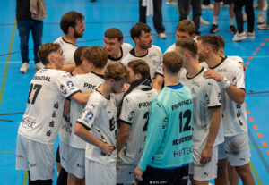 Eine Herren-Handballmannschaft in weißen Trikots sitzt auf einem blauen Hallenplatz zusammen und scheint vor oder während eines Spiels Taktiken zu besprechen.