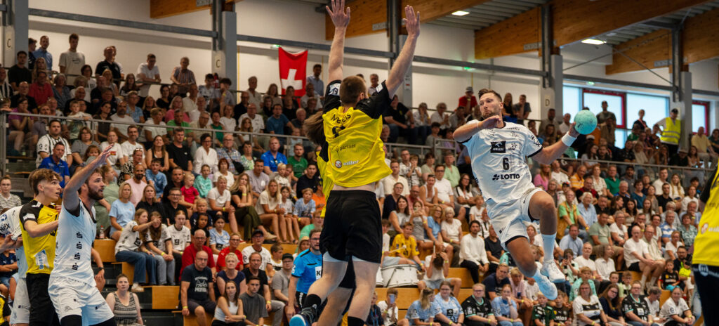 Ein Handballspieler in einem weißen Trikot springt und schießt in Richtung Tor, während ein Verteidiger in einem gelben Trikot aufspringt und den Ball abblockt. Die Zuschauer in der vollbesetzten Sporthalle verfolgen das intensive Spielgeschehen.