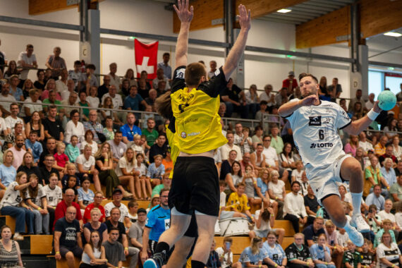 Ein Handballspieler in einem weißen Trikot springt und schießt in Richtung Tor, während ein Verteidiger in einem gelben Trikot aufspringt und den Ball abblockt. Die Zuschauer in der vollbesetzten Sporthalle verfolgen das intensive Spielgeschehen.