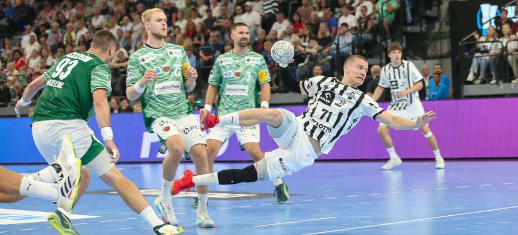 250823_Titel_Supercup_Skipagotu Ein Handballspieler in einem weiß-schwarzen Trikot springt und wirft den Ball in Richtung Tor, umgeben von Verteidigern in grünen Trikots, während eines intensiven Spiels auf einem Hallenplatz mit einer jubelnden Menge im Hintergrund.