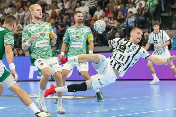 Ein Handballspieler in einem weiß-schwarzen Trikot springt und wirft den Ball in Richtung Tor, umgeben von Verteidigern in grünen Trikots, während eines intensiven Spiels auf einem Hallenplatz mit einer jubelnden Menge im Hintergrund.