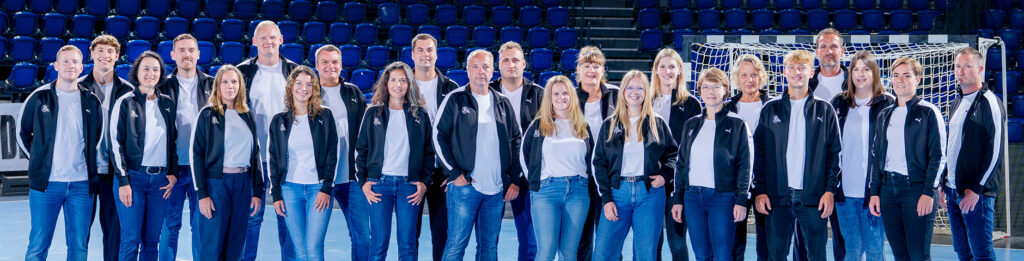 2526_Geschäftsstelle_Titel Eine Gruppe von 22 Personen in passenden schwarzen Jacken und weißen Hemden steht in einer Reihe auf einem Sportplatz mit einem Netztor und leeren blauen Stadionsitzen im Hintergrund.