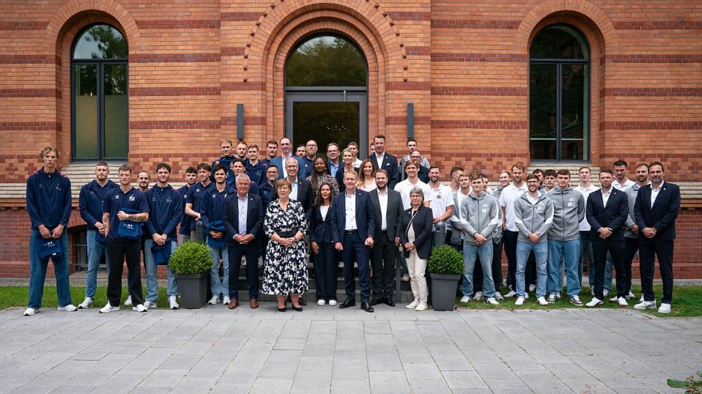 Eine große Gruppe von Menschen, meist Männer in Sportkleidung und einige wenige in formeller Kleidung, posieren für ein Foto vor einem roten Backsteingebäude mit Bogenfenstern und zwei Topfsträuchern auf beiden Seiten.
