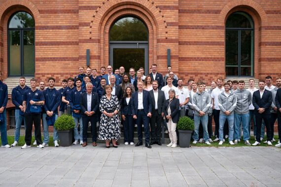 Eine große Gruppe von Menschen, meist Männer in Sportkleidung und einige wenige in formeller Kleidung, posieren für ein Foto vor einem roten Backsteingebäude mit Bogenfenstern und zwei Topfsträuchern auf beiden Seiten.